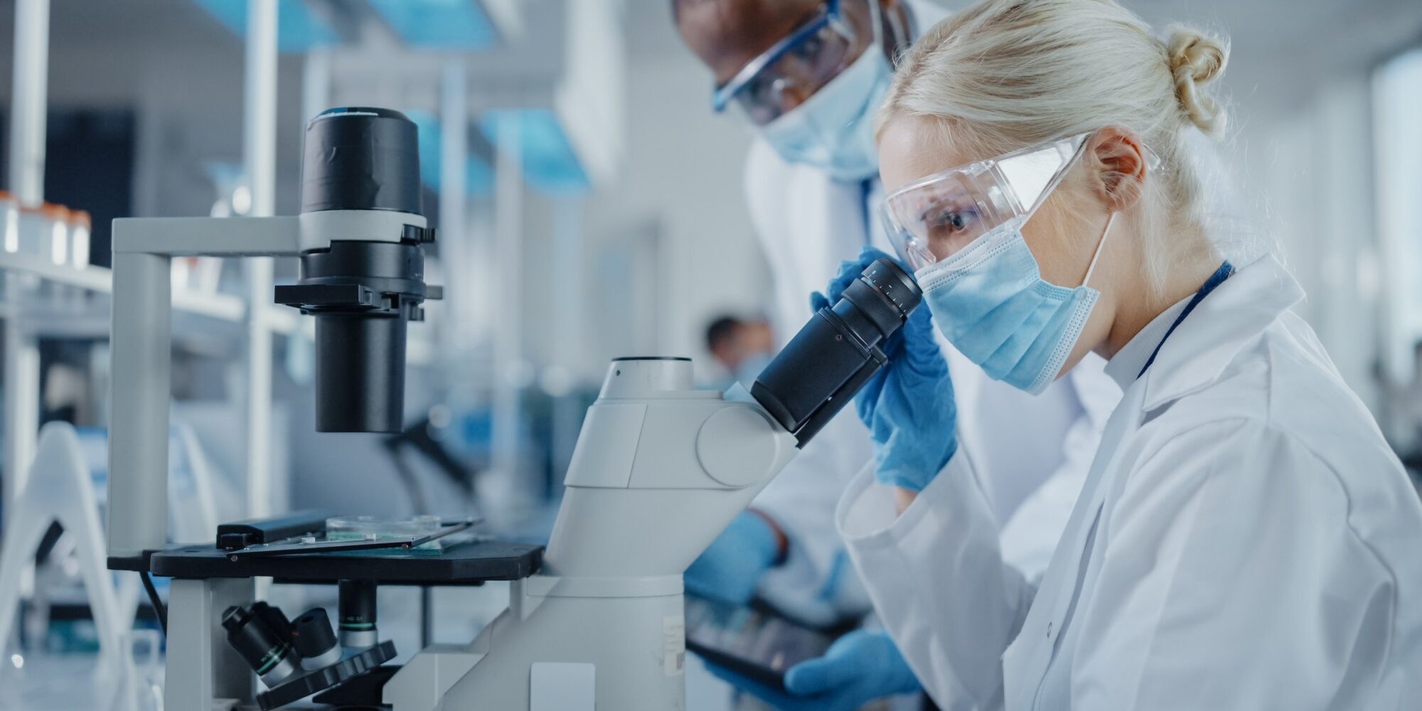 Modern,Medical,Research,Laboratory:,Two,Scientists,Wearing,Face,Masks,Working Modern Medical Research Laboratory: Two Scientists Wearing Face Masks Working Together Using Microscope, Analysing Samples, Talking. Advanced Scientific Lab for Medicine, Biotechnology.